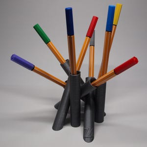 Può includere: Un portapenne nero con più scomparti, contenente penne colorate. Le penne hanno fusti arancioni e cappucci di vari colori, tra cui blu, verde, rosso e giallo. Il portapenne è realizzato in un materiale grigio scuro.