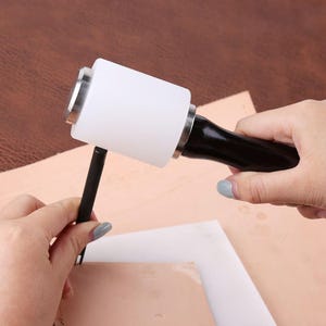 Pode incluir: Um ma&ccedil;o de artesanato em couro branco e prateado com cabo preto &eacute; usado para golpear um pun&ccedil;&atilde;o de metal. O pun&ccedil;&atilde;o &eacute; posicionado em um peda&ccedil;o de couro marrom claro. Ferramentas para trabalhos em couro e artesanato.