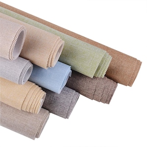 Può includere: Assortimento di tessuti arrotolati in vari colori neutri, tra cui beige, grigio, blu e marrone. Il tessuto ha un aspetto strutturato, che suggerisce un materiale tessuto. I rotoli sono ben disposti, mostrando le diverse sfumature e trame.