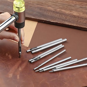 May include: A set of silver metal leather hole punch tools with a yellow and silver mallet. The tools are on a brown leather surface. The tools are used for leathercraft and other crafting projects.