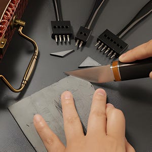 Pode incluir: Uma vista de perto de ferramentas de couro, incluindo uma faca, pun&ccedil;&otilde;es e um peda&ccedil;o de couro. A faca tem uma al&ccedil;a preta com um detalhe dourado. Os pun&ccedil;&otilde;es t&ecirc;m v&aacute;rios dentes. Uma mala de couro marrom com uma al&ccedil;a dourada &eacute; vis&iacute;vel.