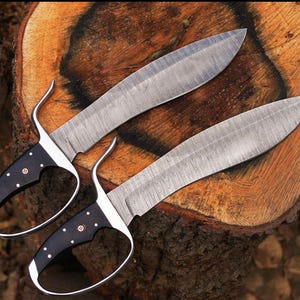 May include: Two Damascus steel knives with black handles and silver accents. The blades have a curved shape and are set against a wooden background. The handles feature white dot and copper rivet details.