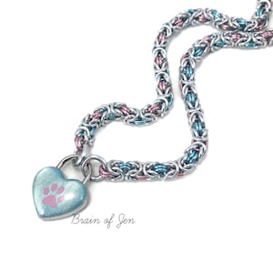 May include: A silver chainmail necklace with a heart-shaped pendant. The pendant is light blue with a pink paw print. The chain is a mix of silver, blue, and pink links. The text "Brain of Jen" is visible.
