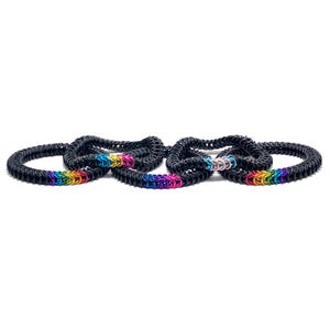 May include: A set of five black chainmail bracelets with rainbow coloured accents. The bracelets are stacked on top of each other.