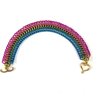 Pode incluir: Uma pulseira colorida de malha de corrente em forma de arco-íris. A pulseira apresenta anéis rosa, dourados e azuis. O fecho é dourado e tem um design em forma de coração. A pulseira está em um fundo branco.