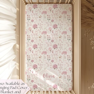 May include: A crib sheet with a floral pattern in shades of pink and green on a cream background. The sheet is inside a wooden crib. The name "Olivia Kate" is printed in a decorative frame on the sheet.