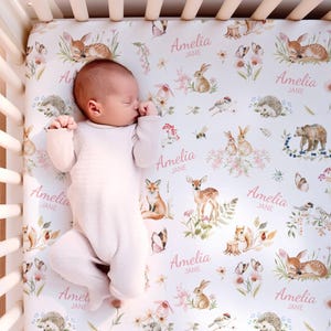 Könnte beinhalten: Ein Baby in einem hellrosa Strampler liegt auf einem Bettlaken mit einem Waldtiermuster. Das Laken zeigt Illustrationen von Hirschen, Kaninchen, Füchsen und Bären sowie den Namen "Amelia Jane" in rosa Schrift. Das Kinderbett ist aus hellem Holz.