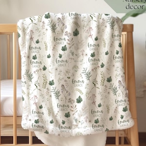 Puede incluir: Una manta de bebé color crema con un estampado botánico y el nombre "Emma Grace" repetido. La manta está sobre una cuna de madera. Las palabras "Botanical Nursery decor" están en la esquina superior derecha.