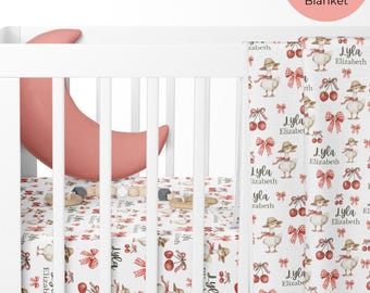 Op de afbeelding: Een witte wieg met een roze halve maan kussen en een set van lakens en dekens. Het laken en de deken hebben een patroon van eenden, kersen en strikken. De tekst "Lyla Elizabeth" wordt herhaald in het ontwerp.