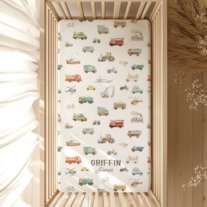 May include: A white fitted crib sheet with a pattern of various vehicles in muted colors, including trucks, trains, and helicopters. The sheet is inside a wooden crib. The name "Griffin James" is printed on the sheet.
