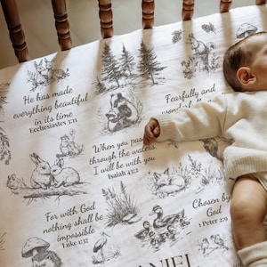 May include: A white baby blanket with a woodland animal design, including rabbits, foxes, and squirrels. The blanket features inspirational quotes from the Bible. The blanket is in a crib with wooden rails.