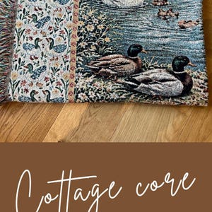 May include: A woven blanket with a pastoral scene of ducks and swans on a lake, with a floral border. The blanket has a fringed edge and is displayed on a wooden surface. The text "Cottage core BLANKET GIFT" is also visible.