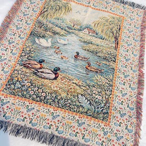 May include: Woven throw blanket featuring a pastoral scene of ducks swimming in a pond, surrounded by lush greenery and trees. The border has a repeating pattern of ducks and flowers, with a fringed edge. The color palette includes blues, greens, and earth tones.