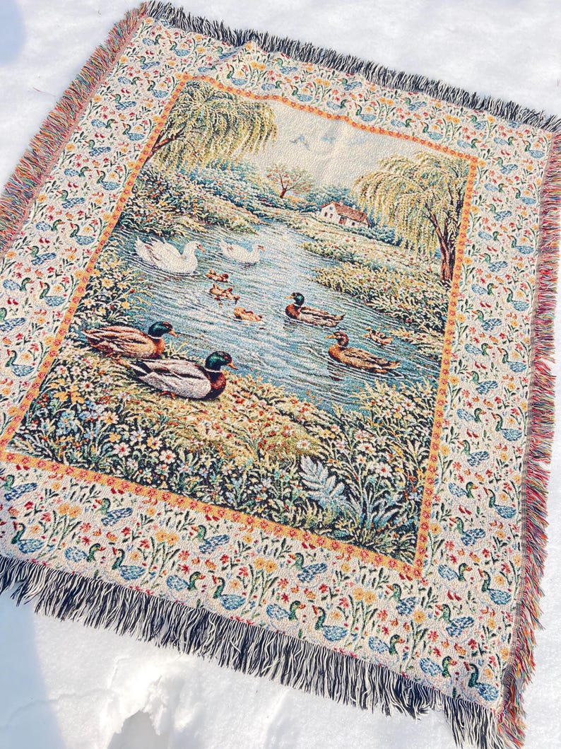 May include: Woven throw blanket featuring a pastoral scene of ducks swimming in a pond, surrounded by lush greenery and trees. The border has a repeating pattern of ducks and flowers, with a fringed edge. The color palette includes blues, greens, and earth tones.