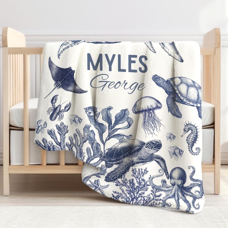 May include: A white baby blanket featuring a blue marine life design, including a stingray, lobster, fish, coral, jellyfish, seahorse, turtle, and octopus. The blanket has the name "MYLES George" printed in blue.