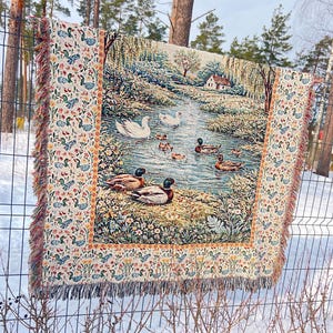 May include: A tapestry throw blanket featuring a serene pond scene with ducks and swans. The border has a repeating pattern of ducks and floral elements. The blanket has a fringed edge and is displayed outdoors on a fence.