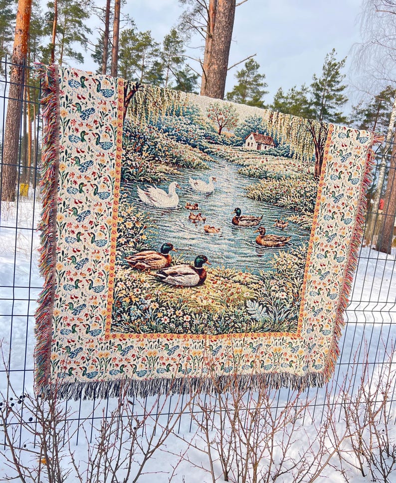 May include: A tapestry throw blanket featuring a serene pond scene with ducks and swans. The border has a repeating pattern of ducks and floral elements. The blanket has a fringed edge and is displayed outdoors on a fence.