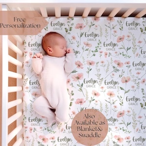 May include: A baby lies on a crib sheet with a floral pattern of pink flowers and green leaves. The sheet is personalized with the name "Evelyn Grace". The image also shows the text "Free Personalization" and "Also Available as Blanket & Swaddle".