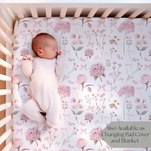 May include: A baby in a light pink onesie lies on a crib sheet with a floral pattern of pink flowers and bows. The crib is white with wooden slats. Text on the sheet reads "Also Available as Changing Pad Cover and Blanket."