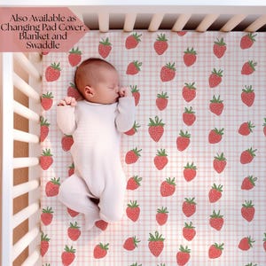 Könnte beinhalten: Ein Baby schläft in einem Kinderbett auf einem rosa-weiß karierten Laken mit Erdbeermuster. Das Kinderbett ist weiß, und das Laken hat rote Erdbeeren mit grünen Stielen. Der Text oben links lautet "Also Available as Changing Pad Cover, Blanket and Swaddle."