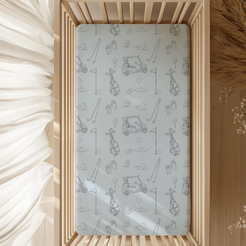 May include: A light blue fitted crib sheet with a repeating pattern of golf-themed illustrations in gray. The design includes golf clubs, golf bags, golf carts, flags, gloves, and golf balls. The sheet is displayed in a wooden crib.
