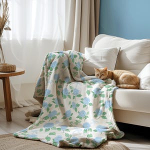 Puede incluir: Una manta crema con un estampado floral de hortensias azules y hojas verdes sobre un sofá blanco. Un gato atigrado naranja descansa en el sofá. La manta añade un toque de color a la sala de estar de tonos neutros.