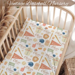 May include: A crib with a baseball-themed nursery set. The set includes a white fitted sheet with baseball bats, gloves, jerseys, and pennants. The sheet has the text "Oliver Elias" and "Let's Play Ball". The top of the image has the text "Vintage Baseball Nursery".
