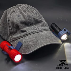 May include: A gray, worn baseball cap with two small clip-on flashlights. One flashlight is red, the other is blue. Both flashlights are turned on, emitting bright white light. The logo "ARCH CITY PRINT FORGE" is visible in the bottom right corner.