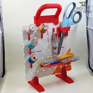 May include: A red and white sewing organizer with a handle, holding spools of thread, scissors, a measuring tape, and other sewing tools. The organizer has a grid-like pattern and a small shelf for accessories.