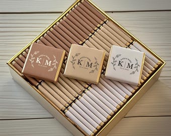 Può includere: Una scatola bordata d'oro piena di barrette di cioccolato marroni e bianche. Sono visibili tre quadratini di cioccolato con le iniziali "KM" e la data "30.11.2024". La scatola è appoggiata su una superficie di legno chiaro.