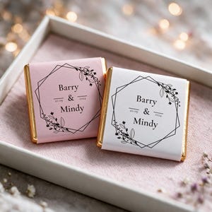 以下が含まれることがあります： 金色のホイルで包まれた2つの正方形のチョコレートバー。紙製のスリーブが付いており、1つはライトピンク、もう1つは白です。どちらも黒い幾何学的なフレーム、花のアクセント、Barry & Mindyという文字と20.09.2024という日付が印刷されています。