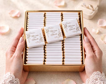 May include: A rectangular gold box filled with rows of small, white-wrapped chocolate squares. Each square features a gold-foiled floral design and the printed text Mariam and Roberth. The box is presented in a clear, organized arrangement.