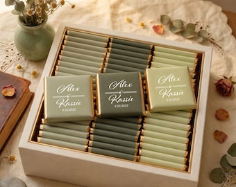 May include: A white wooden box filled with rows of rectangular chocolate bars wrapped in sage green and dark olive paper. Three bars feature white printed text reading Alex and Kassie with the date 4.25.2025 and a small heart icon.