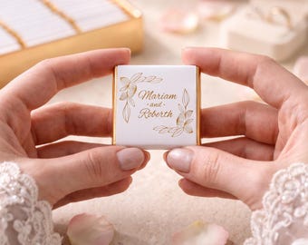 May include: A square white chocolate bar wrapper featuring gold foil-stamped floral corner accents and the text Mariam and Roberth printed in the center. The wrapper has a metallic gold edge and is held between two hands with lace sleeves.