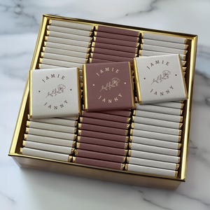 Könnte beinhalten: Eine goldene Schachtel gefüllt mit einzeln verpackten Schokoladentafeln. Die Schokoladentafeln sind mit einem floralen Design und den Namen "Jamie" und "Janny" verziert.