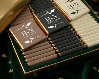 May include: A gold box containing chocolate bars in shades of brown, black, and white. Each bar is wrapped in gold foil and has a custom label with the initials "H N", the names "Hailey Nelson", and the date 05.04.2023. The box sits on a green velvet surface.