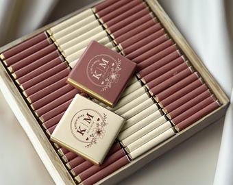 Può includere: Una scatola di legno piena di cioccolatini confezionati singolarmente. I cioccolatini sono disposti in file di colori alternati, alcuni in una tonalità rosa polveroso e altri in crema. Due cioccolatini in cima presentano le iniziali "KM" e disegni floreali.