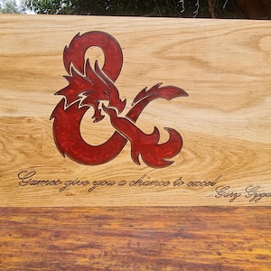May include: Wooden tabletop gaming screen with a red dragon and ampersand design. The screen is made of light brown wood and features the text "Games give you a chance to excel" and the signature "Gary Gygax".