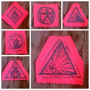 May include: A collection of six pink fabric patches with dark purple graphic designs. The patches feature various symbols, including a flame, an eye, a running figure with the text "GET LOST", and geometric shapes. The patches have a raw, unfinished edge.