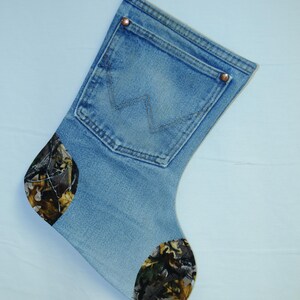 May include: A denim Christmas stocking with a pocket and copper rivets. The toe and heel are patterned with a camouflage design. The stocking is hanging from a white hook.