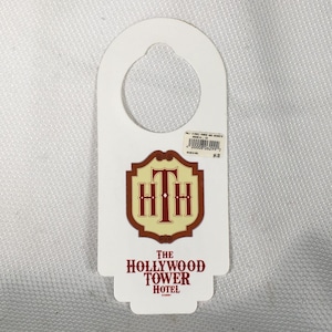 May include: White door hanger with a circular cutout at the top. The design features a maroon and gold emblem with the letters "HTH" and the words "The Hollywood Tower Hotel" in maroon text.