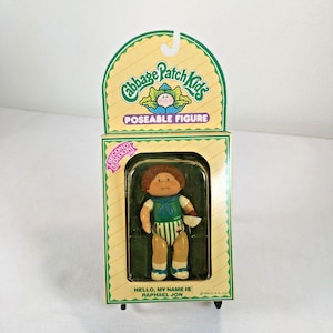 May include: A Cabbage Patch Kids poseable figure in its original packaging. The doll has brown hair and wears a green and white striped outfit. The packaging features the text Cabbage Patch Kids, Poseable Figure, Second Edition, and Hello, My Name Is Raphael Jon.
