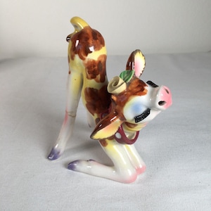 Op de afbeelding: Een vintage keramieken beeldje van een knielend hert met een bloemenhoofdtooi. Het hert heeft bruine vlekken, een roze neus en blauwe ogen. Het beeldje is beschilderd met geel, bruin en wit.