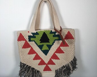 Op de afbeelding: Crèmekleurige draagtas met een geometrisch ontwerp in rood, groen en zwart. De tas heeft een geweven textuur, crèmekleurige handvatten en zwarte franjes aan de onderkant. Een bruin leren label hangt aan de zijkant.