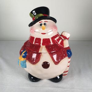 May include: A ceramic snowman cookie jar with a black top hat, red scarf, and a blue gift. The snowman has a smiling face with rosy cheeks and a carrot nose. The jar is a festive holiday decoration.