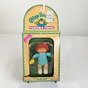 May include: A Cabbage Patch Kids poseable figure in its original packaging. The doll has red hair, a light blue outfit, and white socks. The box features the text Cabbage Patch Kids, Poseable Figure, First Edition, and Hello, My Name Is Joanie Ann.
