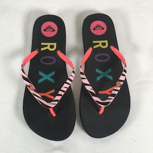 May include: Pair of black Roxy flip-flops with pink and zebra-striped straps. The flip-flops have the Roxy logo in a circle at the top and the word "ROXY" in colorful letters. The flip-flops are on a white background.
