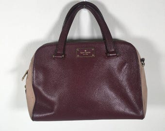 Op de afbeelding: Een bordeauxrode en beige leren satchel handtas met dubbele handvatten. De tas heeft een getextureerde buitenkant, een goudkleurige logoplaat en een ritssluiting. De tas is een Kate Spade-ontwerp.