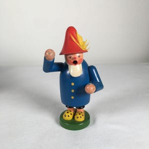 May include: A wooden figurine of a man in a blue coat with yellow buttons, a red hat with a yellow feather, and yellow shoes with black spots. The figurine is standing on a green base.