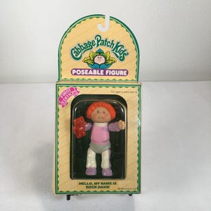 Puede incluir: Una figura articulada de Cabbage Patch Kids en su embalaje original. La muñeca tiene el pelo naranja brillante, una camiseta rosa de manga larga y pantalones grises, y sostiene un pequeño oso rojo. La caja incluye el texto Cabbage Patch Kids, First Edition y Hello, My Name Is Rock David.
